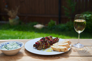 Greek style souvlaki skewer from lamb meat with onion and sweet red pepper served outside in garden with rustic bread, glass of white wine and traditional tzatziki cucumber salad. Barbeque party food.