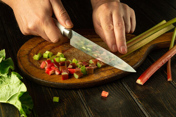 Preparing a vegetarian rhubarb dish on the kitchen table. Chef hands cut the stems of a rheum plant with a knife on a kitchen board before preparing breakfast