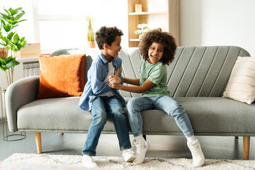 Happy two African American kids having fun while fooling around together, sitting on sofa at home
