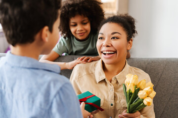 Overjoyed young mother and her kids sitting on couch, holding birthday presents at home
