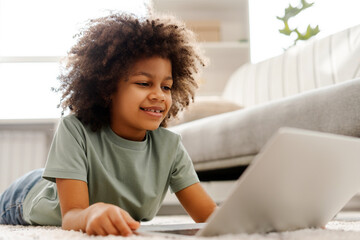 Smiling African American girl lying on the floor in the room at home, using laptop, watching video