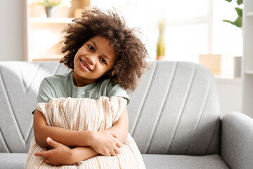 Happy African American little girl sitting on cozy sofa while holding pillow, looking at camera