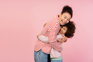 Happy young mother hugging her little daughter while standingin studio, isolated on pink background