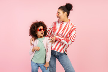 Surprised young woman and little girl in stylish sunglasses posing in studio, isolated on pink