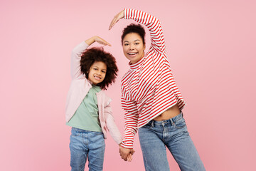 Smiling mother and her daughter posing in studio and making heart by their hands, isolated on pink