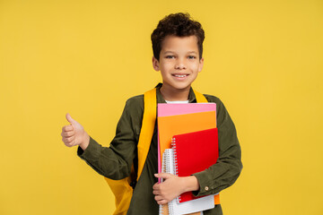 African American boy looking at camera, holding books, with backpack, isolated on yellow background