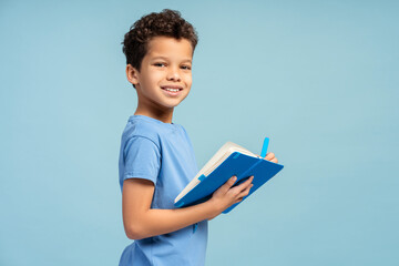 Happy little boy writing in notebook, looking at camera. Back to school concept