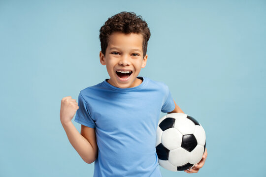 Portrait of emotional child boy holding soccer ball in his hand, isolated on blue background