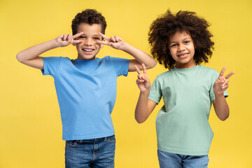 Happy little cheerful friends showing v-sign while posing in studio, isolated on yellow background
