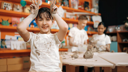 Asian girl pose at camera while diverse children modeling clay behind. Happy cute student wearing...