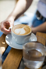 A cup of cappuccino with heart shaped foam being held by woman's hand on a wooden table