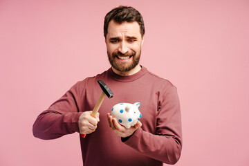 Scared bearded man holding iron and broking piggy bank, isolated over pink studio background