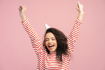 Excited girl, teenager wearing birthday cone rejoicing with raised arms and closed eyes