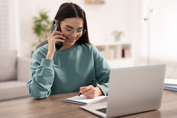 Fototapeta premium Young woman talking on smartphone during webinar at table in room