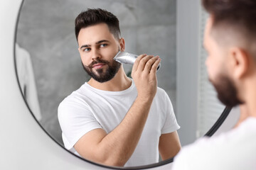 Handsome young man trimming beard near mirror in bathroom