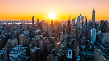 Orange light of setting sun illuminating the skyscrapers and high-rise buildings in New York scenery. Top view on the metropolis from above the East River. - obrazy, fototapety, plakaty