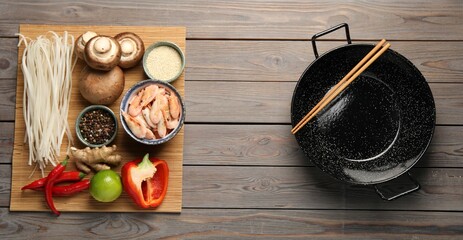 Black wok, chopsticks and bamboo mat with products on color wooden table, top view