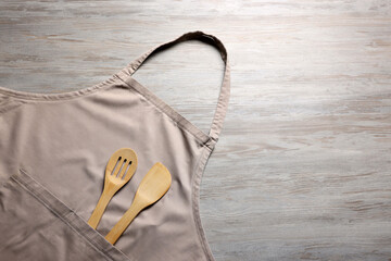 Stylish beige apron, spatula and slotted spoon on light wooden table, top view. Space for text