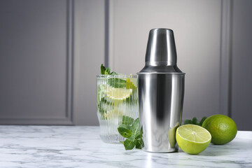 Metal cocktail shaker, delicious mojito, limes and mint on white marble table, space for text