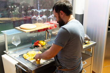 A man cutting potatoes on a cutting board in a kitchen. There are several wine glasses on the...