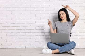 Student with laptop sitting on floor and pointing at something near white brick wall. Space for text