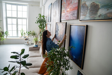 Woman painter hanging newly painted modern framed painting on wall in bright room art workshop with...