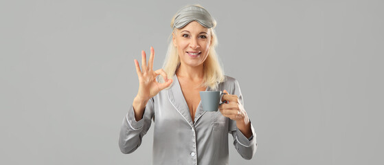 Mature woman in pajamas with cup of coffee showing OK gesture on grey background