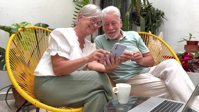 Senior adult couple sitting on home balcony relaxing with a coffee cup, retired seniors looking together at the same mobile phone smiling. Retirement lifestyle concept
