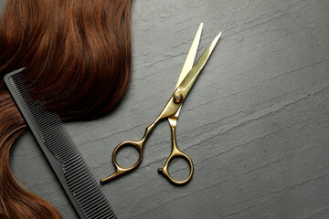 Professional hairdresser scissors and comb with brown hair strand on dark grey table, top view....