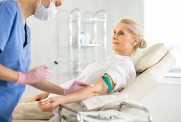 Smiling relaxed elderly female patient sitting on couch in medical office, getting vitamin therapy. Professional medical worker administering intravenous injection