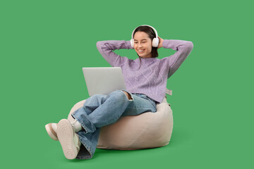Beautiful young happy woman in headphones with laptop resting on beanbag chair against green background