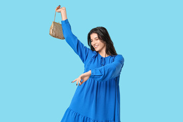 Beautiful young woman in stylish blue dress with chain bag dancing on color background