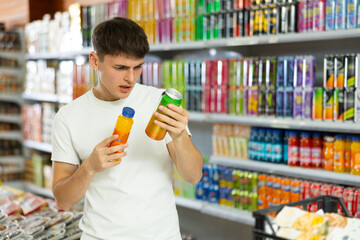 Thoughtful young guy choosing soft drinks in grocery store, standing by product shelves and reading...