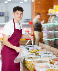 Young guy in supermarket, scooping marinated olives from bucket with ladle