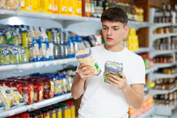 Portrait of pensive young guy choosing organic dried vegetables in supermarket, holding packages...