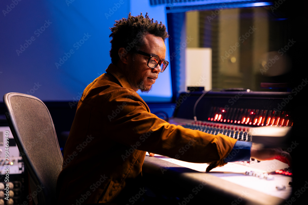 Poster african american music producer operates audio console with moving faders and meters, recording trac