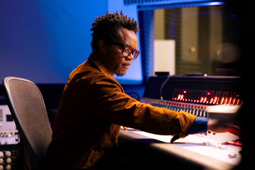 African american music producer operates audio console with moving faders and meters, recording...