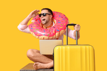 Happy office worker in sunglasses with inflatable ring, laptop and suitcase sitting on table against yellow background