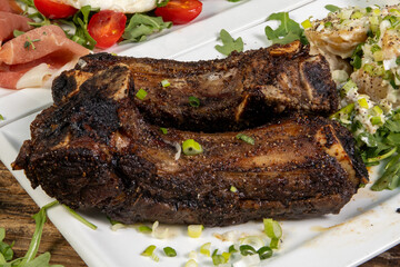 Big juicy smoked meaty BBQ beef ribs with a baked potato with sour cream and chives and smoked ham and tomatoes in the background
