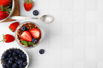 Tasty granola, strawberries, blueberries and spoon on white tiled table, flat lay. Space for text