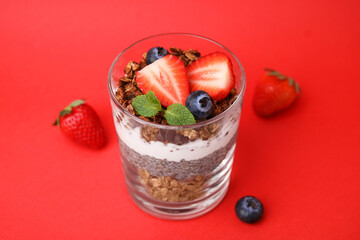 Tasty granola with berries, yogurt and chia seeds in glass on red background, closeup