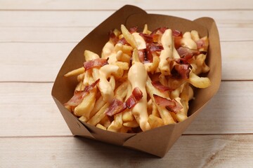 Tasty potato fries, cheese sauce and bacon in paper container on light wooden table, closeup