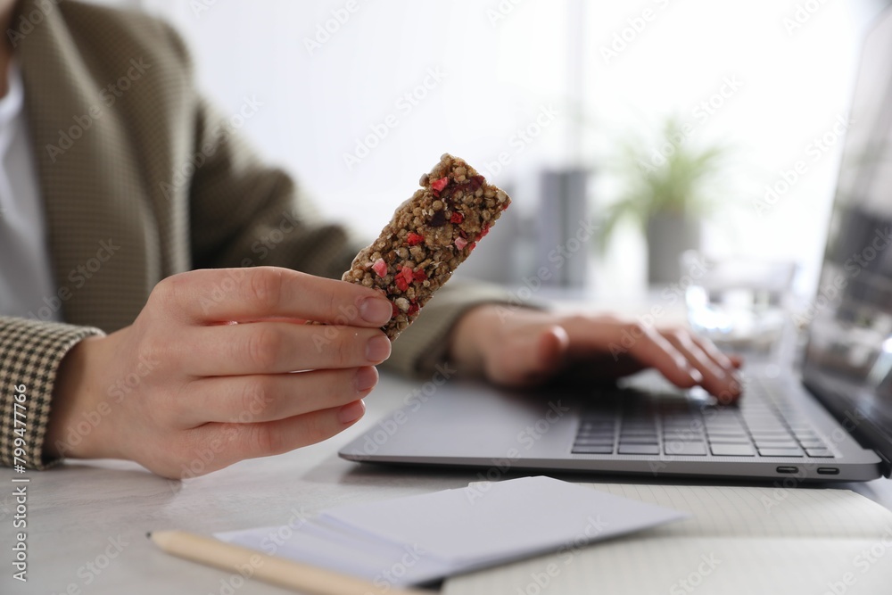 Wall mural Woman holding tasty granola bar working with laptop at light table in office, closeup