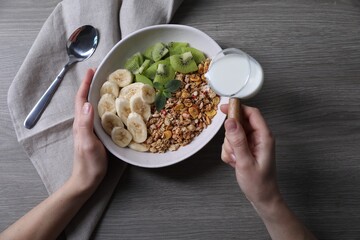 Woman pouring milk into bowl of tasty granola with banana and kiwi at grey wooden table, top view