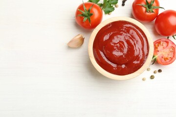Delicious ketchup in bowl, peppercorns and tomatoes on white wooden table, flat lay. Space for text