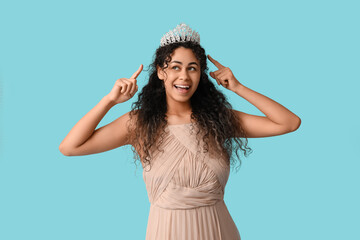 Beautiful young African-American woman in stylish prom dress pointing at tiara on blue background