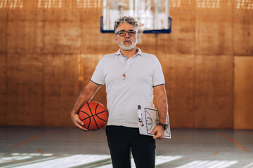 Mature confident male coach holding basketball ball and a coach clipboard