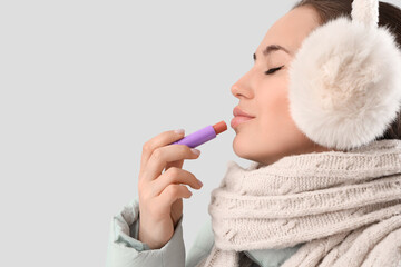 Young woman in winter clothes applying lip balm on light background, closeup