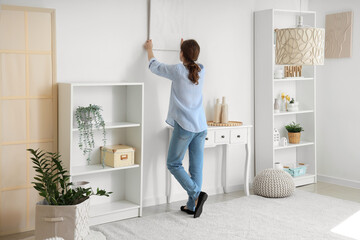 Young pretty woman hanging picture on wall in white room