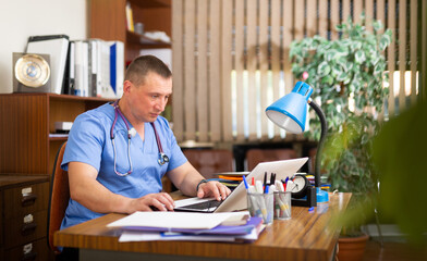 Qualified middle aged man doctor sitting in office at workplace and working at laptop